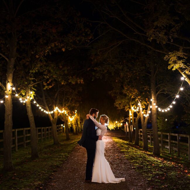 Newly married couple standing of the drive at Wood Farm at Everdon in the evening. There are lights lining the driveway and lights behinds them.