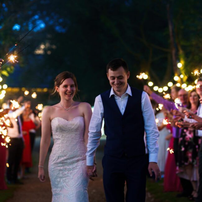 Couple celebrating at their wedding reception at Wood Farm Everdon. Couple are smiling, walking through a line of their friends who are all holding sparklers.